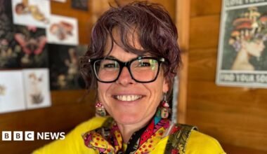 Kylie Dixon is pictured seated inside her allotment shed. She is wearing dark rimmed glasses, a yellow t-shirt and green dungarees with a brightly coloured neck-scarf and mushroom-shaped earrings. She has purple-tinted dark curly hair that is tied up.