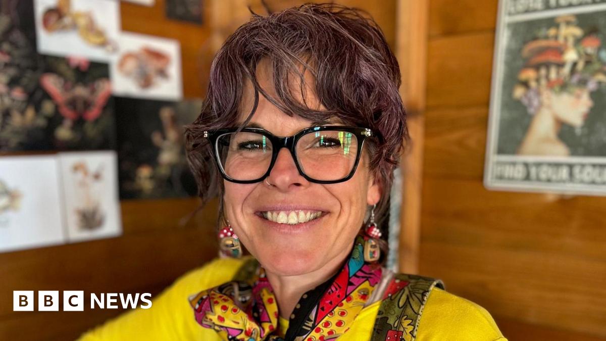 Kylie Dixon is pictured seated inside her allotment shed. She is wearing dark rimmed glasses, a yellow t-shirt and green dungarees with a brightly coloured neck-scarf and mushroom-shaped earrings. She has purple-tinted dark curly hair that is tied up.