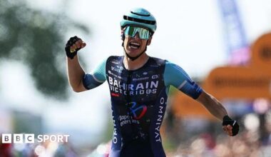 Alec Segaert celebrates by appearing to punch the air, while wearing Bahrain-Victorious cycling outfit, a helmet and sunglasses