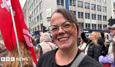A woman with long brown hair and clear-lensed glasses wears red lipstick as she smiles to the camera. She holds a red flag in her left hand. Behind her his a large crowd of people, out-of-focus.
