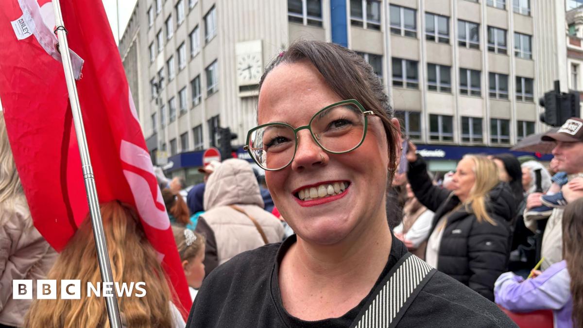A woman with long brown hair and clear-lensed glasses wears red lipstick as she smiles to the camera. She holds a red flag in her left hand. Behind her his a large crowd of people, out-of-focus.