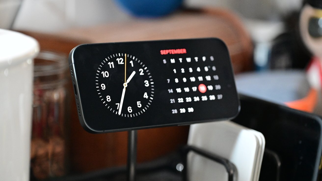 Smartphone mounted horizontally showing a dark analog clock on the left and a September calendar on the right, sitting among kitchen items on a counter