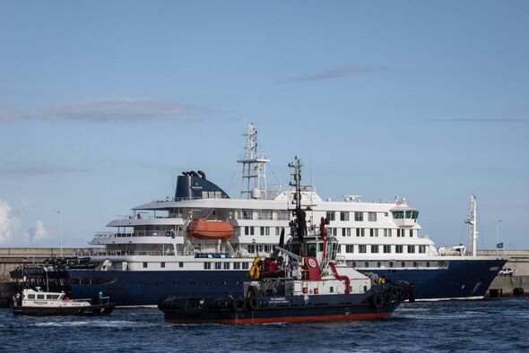 An image of a large cruise ship with multiple decks is seen docked at a port. Nearby, a smaller tugboat is positioned adjacent t An image of a large cruise ship with multiple decks is seen docked at a port. Nearby, a smaller tugboat is positioned adjacent t