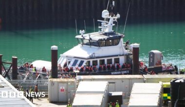 A ship is docked at a port. It is full of people. Most of them are wearing orange life jackets.