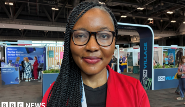 Faridat Ibidun is a black Nigerian woman with long braided black hair worn down over her right shoulder. She is wearing glasses, a black top and red suit jacket. You can see various stalls behind her in a warehouse interior for the RCN's annual congress.