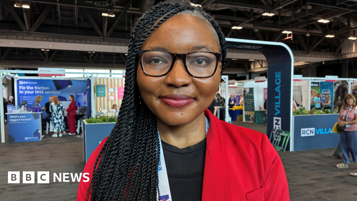 Faridat Ibidun is a black Nigerian woman with long braided black hair worn down over her right shoulder. She is wearing glasses, a black top and red suit jacket. You can see various stalls behind her in a warehouse interior for the RCN's annual congress.