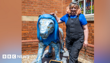 Sam Morris standing next to Kintsugi the lion. She is wearing black dungarees and a blue t shirt and standing in front of a brick building.