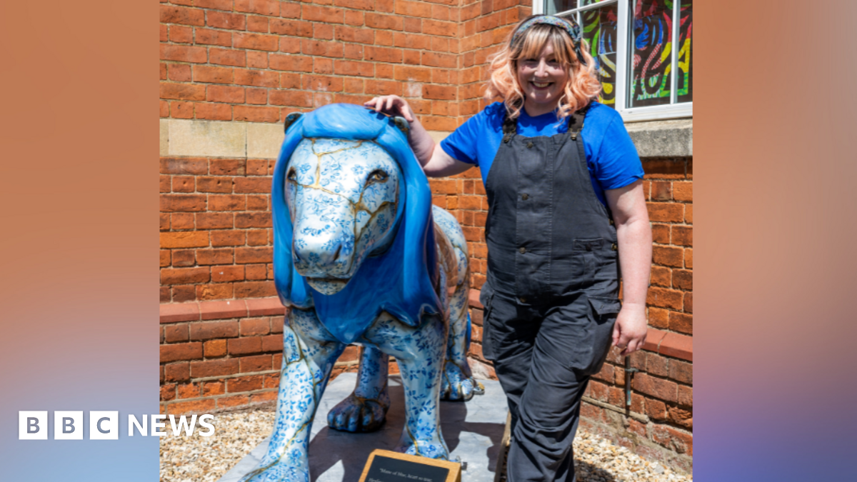 Sam Morris standing next to Kintsugi the lion. She is wearing black dungarees and a blue t shirt and standing in front of a brick building.