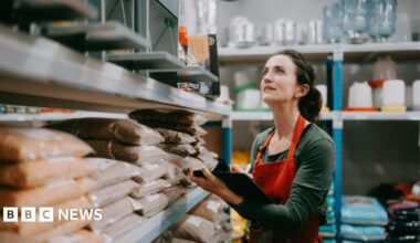Woman working in warehouse - she is wearing a red apron and green top and carrying a clipboard as she stands by shelves