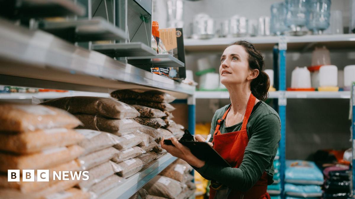 Woman working in warehouse - she is wearing a red apron and green top and carrying a clipboard as she stands by shelves