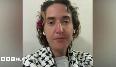 A woman with short hair sitting looking at the camera. She is wearing a keffiyeh, has earbuds in and has a flower above her ear.
