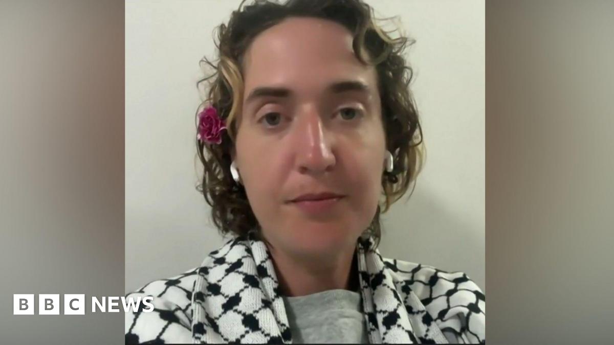 A woman with short hair sitting looking at the camera. She is wearing a keffiyeh, has earbuds in and has a flower above her ear.