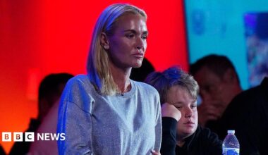 A woman with blonde hair looks ahead seriously. Her hair is all pulled to the right side of her shoulder. She is wearing a silver sparkly sweater and a pendant necklace. There are people in the background.
