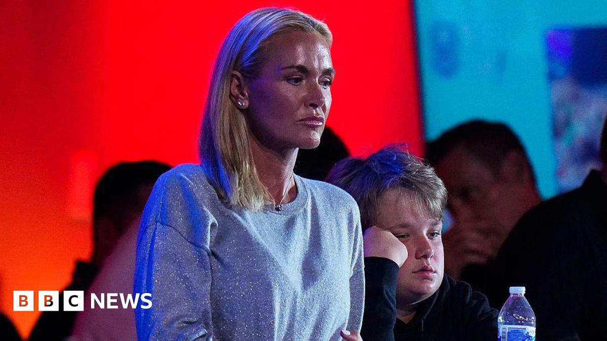 A woman with blonde hair looks ahead seriously. Her hair is all pulled to the right side of her shoulder. She is wearing a silver sparkly sweater and a pendant necklace. There are people in the background.