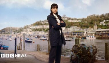 Zahra Ahmadi as the character Det Sgt Esther Williams posing for a press shot with Looe behind her across the water on which yachts are moored. She has long dark hair and is wearing a navy anorak and trousers with a white shirt. She is smiling and has her arms crossed. There are lobster pots at her feet.