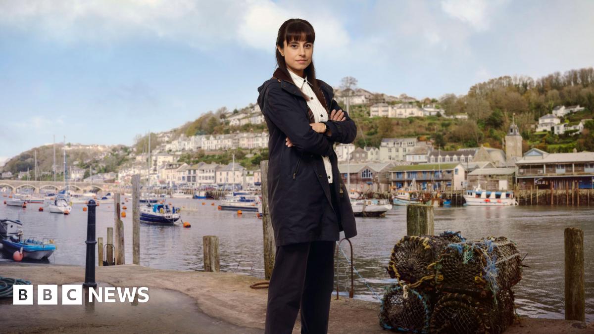 Zahra Ahmadi as the character Det Sgt Esther Williams posing for a press shot with Looe behind her across the water on which yachts are moored. She has long dark hair and is wearing a navy anorak and trousers with a white shirt. She is smiling and has her arms crossed. There are lobster pots at her feet.
