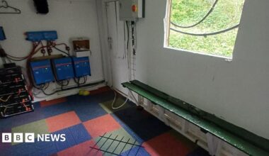 A room with a broken window and a wooden board, which has fallen on a chequered carpet.
