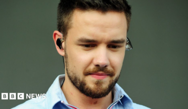 Liam Payne, with dark hair and a beard, is looking down and wearing an earpiece in front of a plain background.