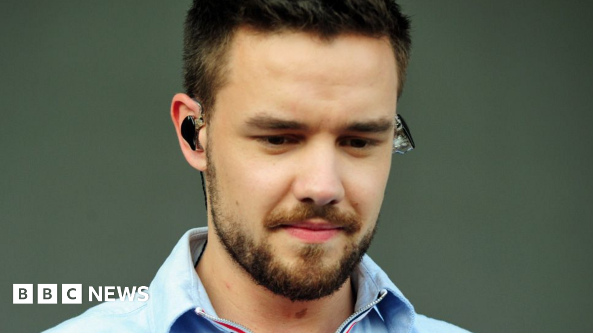 Liam Payne, with dark hair and a beard, is looking down and wearing an earpiece in front of a plain background.