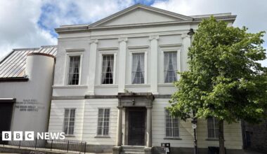 The former Newry Arts Centre - a cream-coloured two-storey building with a replica classical facade and a modern side extension.  The main structure is symmetrical, with five large Georgian-style sash windows on the top floor, intersected by four decorative pilasters.  The front door is protected by a metal grill and sheltered by a pillared granite portico. A disabled access ramp leads to the modern side extension which has a curved exterior wall and a steep roof.