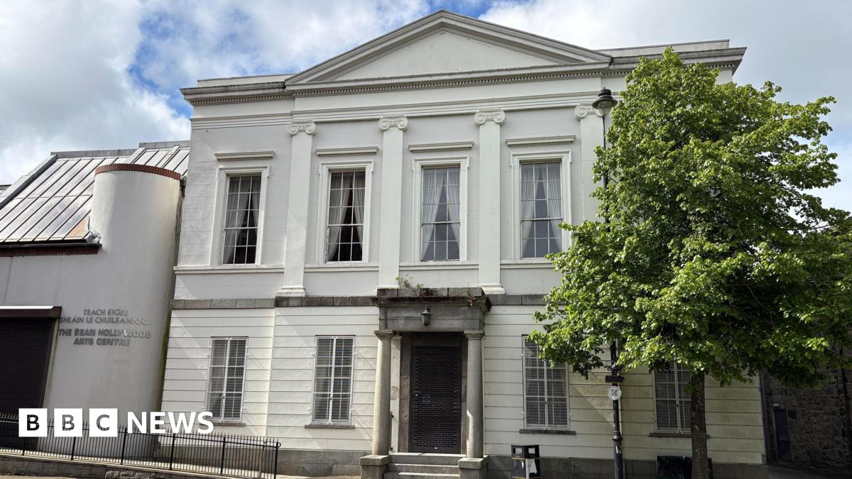The former Newry Arts Centre - a cream-coloured two-storey building with a replica classical facade and a modern side extension.  The main structure is symmetrical, with five large Georgian-style sash windows on the top floor, intersected by four decorative pilasters.  The front door is protected by a metal grill and sheltered by a pillared granite portico. A disabled access ramp leads to the modern side extension which has a curved exterior wall and a steep roof.