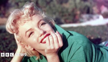 Marilyn Monroe with short blonde hair, wearing a green jacket, resting her chin on her hand outdoors with soft background blur.