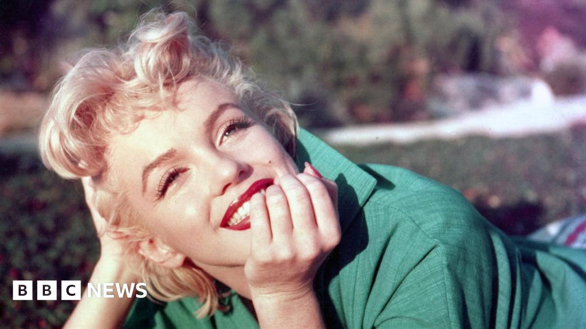 Marilyn Monroe with short blonde hair, wearing a green jacket, resting her chin on her hand outdoors with soft background blur.