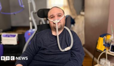 Man in his thirties with short dark hair and stubble, wearing a dark blue jumper looks at camera. He is sitting in a powered wheelchair with a ventilation tube attached to his nose. In the background is medical and hoisting equipment.