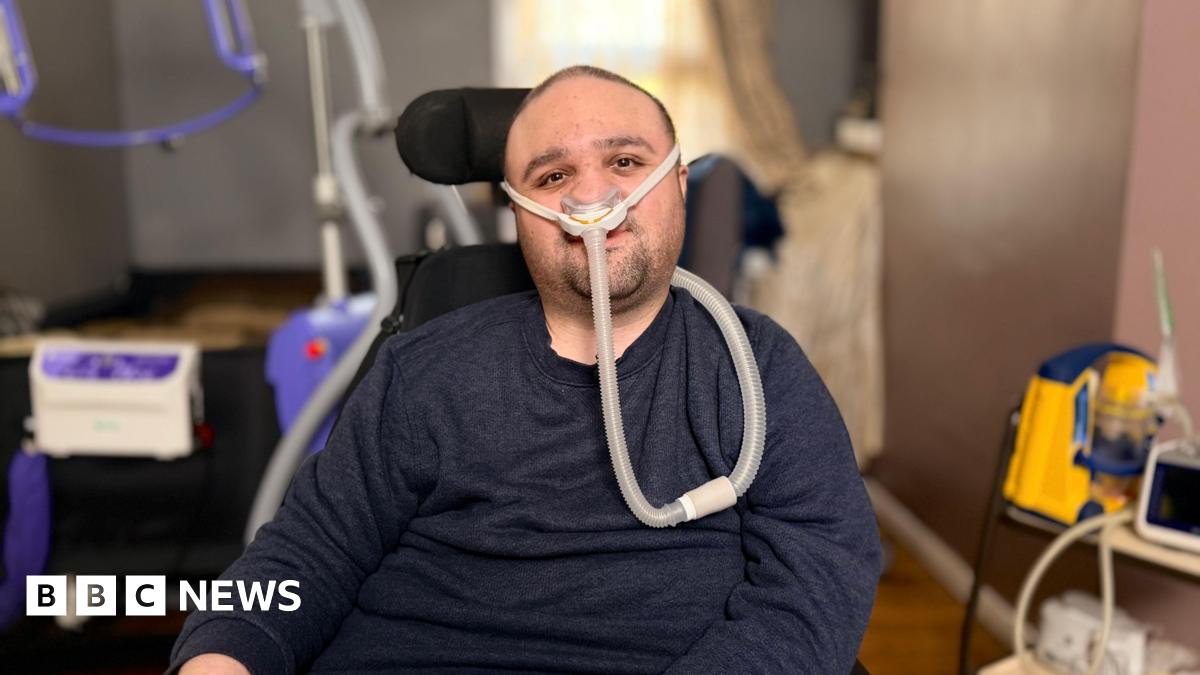 Man in his thirties with short dark hair and stubble, wearing a dark blue jumper looks at camera. He is sitting in a powered wheelchair with a ventilation tube attached to his nose. In the background is medical and hoisting equipment.
