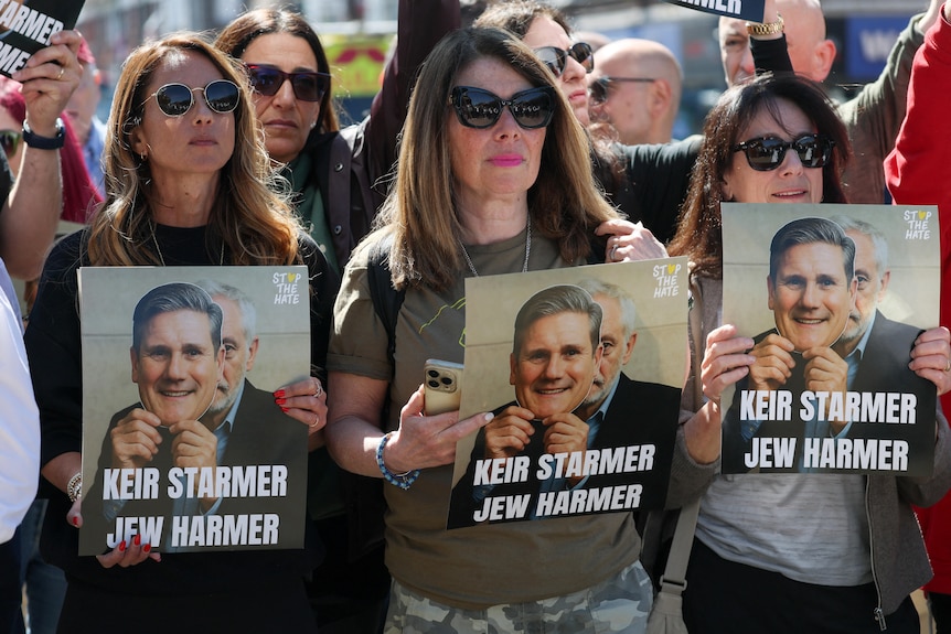 Women stand in a crowd holding pictures depicting Keir Starmer with the words 'Keir Starmer Jew Harmer' on them