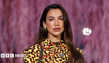 Dua Lipa poses with her dark brown hair straightened and out, while wearing a yellow, black and red Chanel jacket and a gold spiky earring.