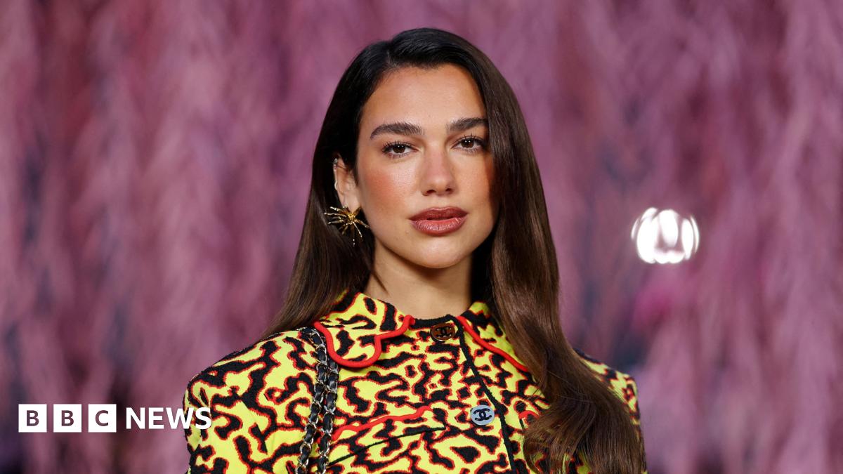 Dua Lipa poses with her dark brown hair straightened and out, while wearing a yellow, black and red Chanel jacket and a gold spiky earring.