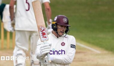 Northamptonshire batter George Bartlett, wearing white gloves, a white shirt and a maroon helmet, raises his bat