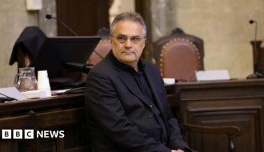 Egisto Ott wearing a dark jacket and black shirt and spectacles, with his hair brushed back looks at the camera as he sits in front of a dark wood-pandelled desk, where the back of two brown leather upholstered chairs can be seen as well as two microphones.