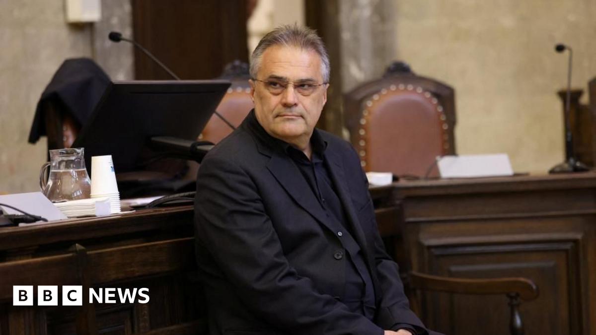 Egisto Ott wearing a dark jacket and black shirt and spectacles, with his hair brushed back looks at the camera as he sits in front of a dark wood-pandelled desk, where the back of two brown leather upholstered chairs can be seen as well as two microphones.