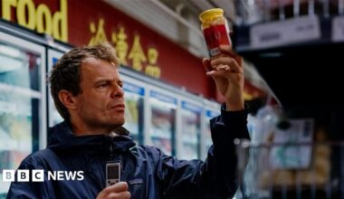 Dan Kelly holding up a jar of pickles and holding a dictaphone in his other hand. Some supermarket fridges are seen in the background.