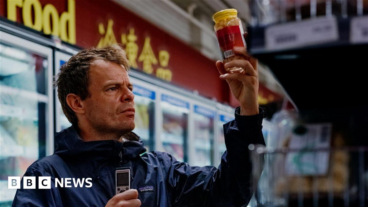 Dan Kelly holding up a jar of pickles and holding a dictaphone in his other hand. Some supermarket fridges are seen in the background.