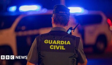 A policeman speaks into a phone with his back turned to the camera, with 'Guardia Civil' written on the back of his vest. A police car with flashing blue lights is visible in the background.