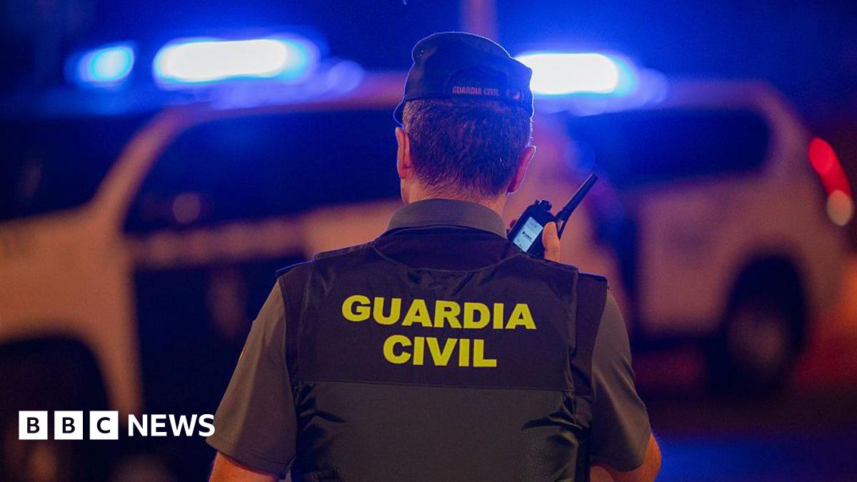 A policeman speaks into a phone with his back turned to the camera, with 'Guardia Civil' written on the back of his vest. A police car with flashing blue lights is visible in the background.