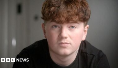 Close-up of Alex's face in the foreground, with short curly ginger hair and a dark top, looking towards the camera. A small neck tattoo is visible above the collar. The background is an indoor setting with a plain wall and a blurred door handle or fixture on the left. No bags, bins, or visible markers are present.