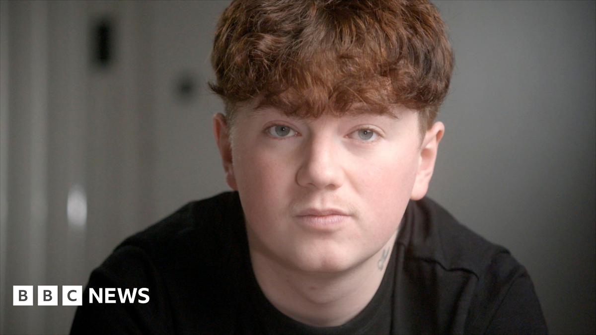 Close-up of Alex's face in the foreground, with short curly ginger hair and a dark top, looking towards the camera. A small neck tattoo is visible above the collar. The background is an indoor setting with a plain wall and a blurred door handle or fixture on the left. No bags, bins, or visible markers are present.