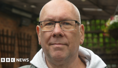 A face-only image of a man wearing a grey hooded jumper and glasses is standing in an outdoor garden area