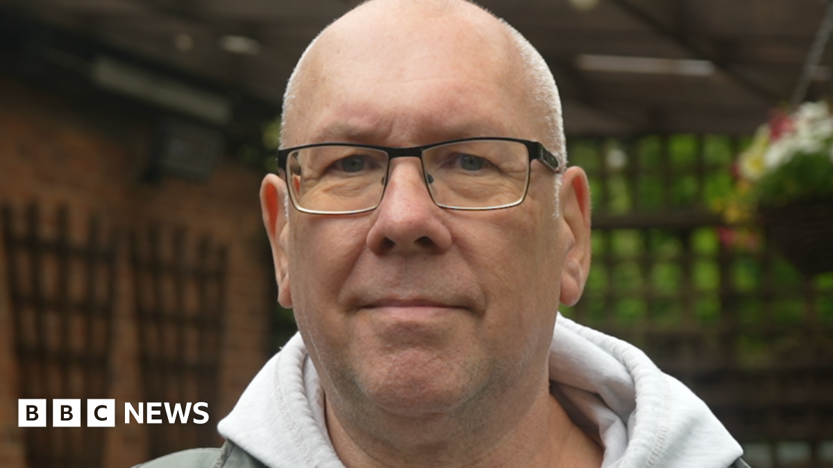A face-only image of a man wearing a grey hooded jumper and glasses is standing in an outdoor garden area