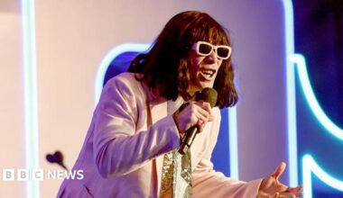 Mr Fantasy performs onstage, wearing pastel pink suit and matching sunglasses. He wears a green scarf, bright pink rings and a short brown wig with a fringe