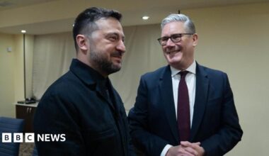 Sir Keir Starmer is stood on the right and is smiling as he looks at Ukrainian President Volodymyr Zelensky. The two men are stood in a hotel conference room which has white walls. Sir Keir is wearing a dark coloured suit jacket, a white shirt, a dark red tie and glasses. Zelensky is dressed in black.