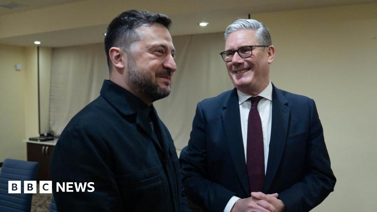 Sir Keir Starmer is stood on the right and is smiling as he looks at Ukrainian President Volodymyr Zelensky. The two men are stood in a hotel conference room which has white walls. Sir Keir is wearing a dark coloured suit jacket, a white shirt, a dark red tie and glasses. Zelensky is dressed in black.