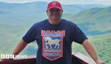 A man stands on a balcony in front of a large expanse of trees and hills. He wears a navy T-shirt with the Ipswich Town logo on it as well as a red cap.