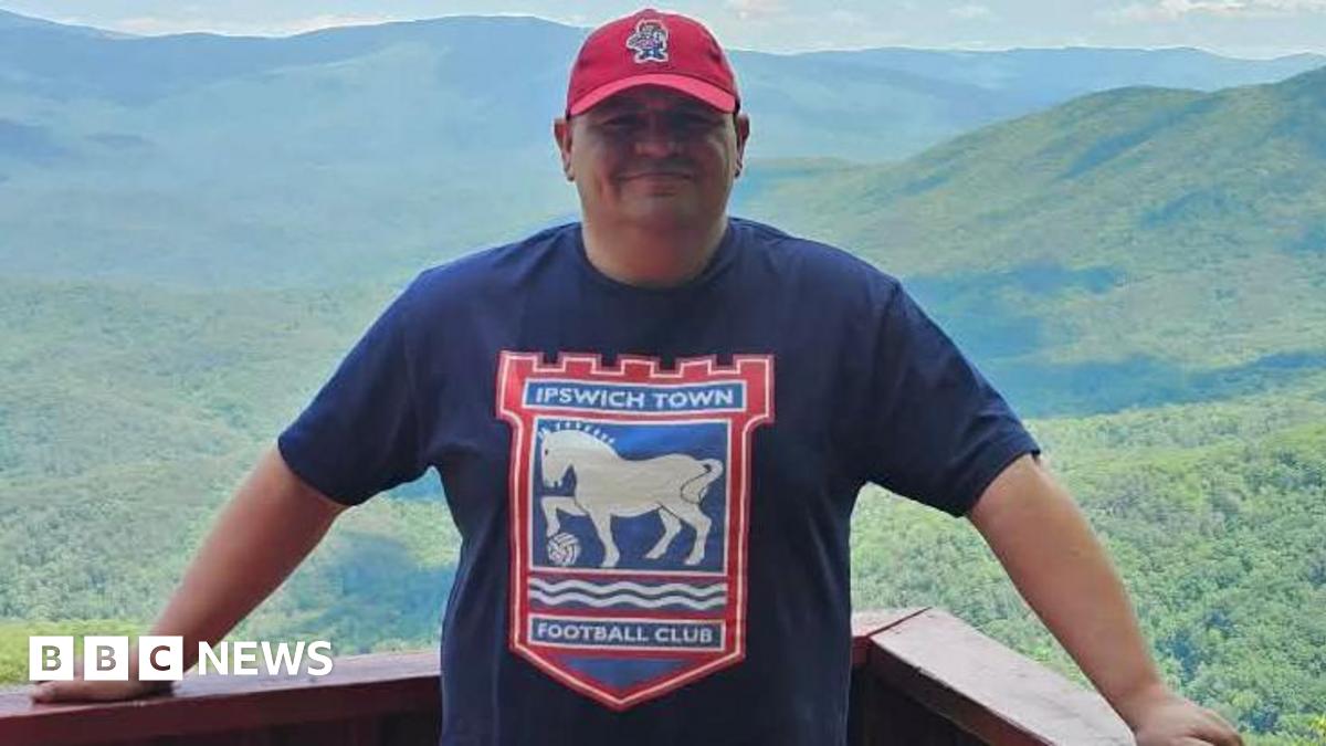 A man stands on a balcony in front of a large expanse of trees and hills. He wears a navy T-shirt with the Ipswich Town logo on it as well as a red cap.