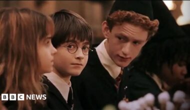 A shot from one of the early Harry Potter films, showing Hermione Granger sitting by Harry Potter and Percy Weasley. Harry looks at the camera directly from side on while Percy speaks to them both, wearing a black cap. Hermione has a fringe and brown shoulder length hair, Harry has a short bowl hair cut and wears school uniform, as does Percy who has reddish wavy short hair.