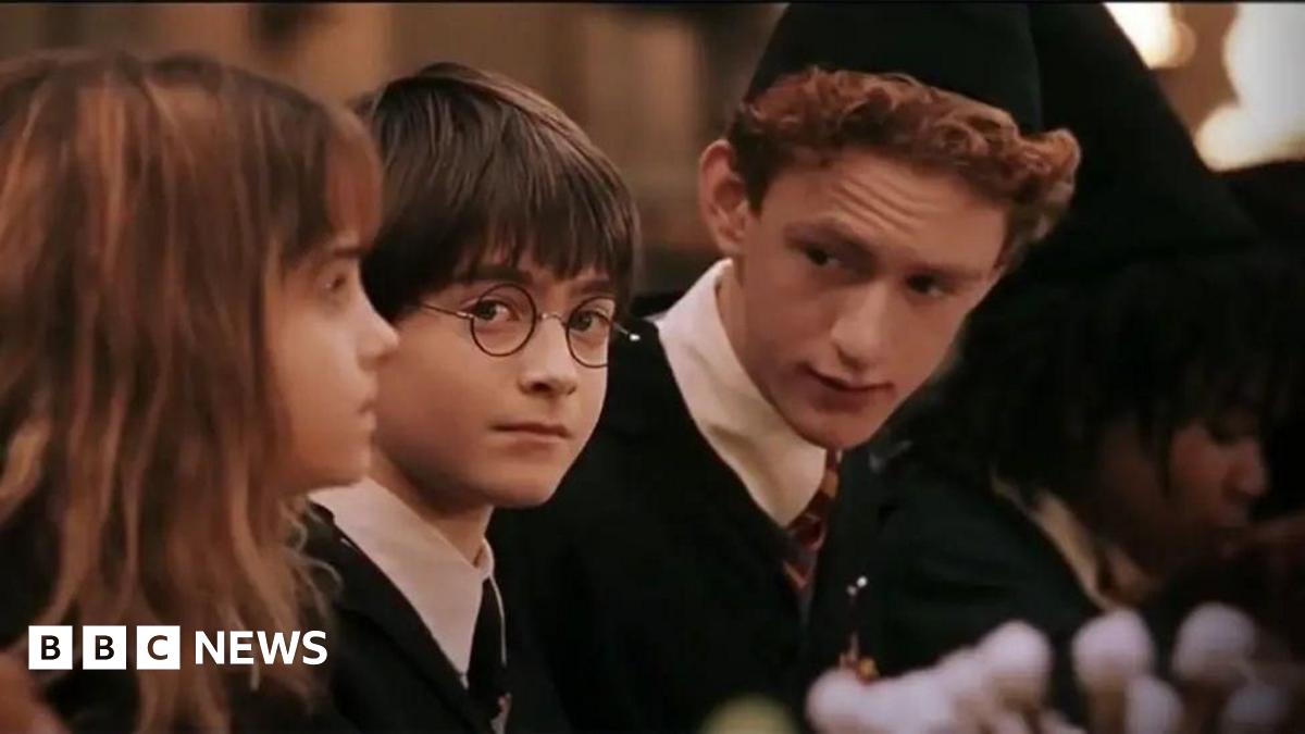 A shot from one of the early Harry Potter films, showing Hermione Granger sitting by Harry Potter and Percy Weasley. Harry looks at the camera directly from side on while Percy speaks to them both, wearing a black cap. Hermione has a fringe and brown shoulder length hair, Harry has a short bowl hair cut and wears school uniform, as does Percy who has reddish wavy short hair.